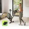 A woman is exercising on a stationary bike in a bright, modern room with large windows overlooking a garden. Produktbild-Vorschau