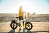 A woman stands on a beach path with a white electric bicycle next to her. The sun is setting, casting a warm glow over the scene. Produktbild-Vorschau