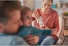 A woman is using a breast pump while sitting in a chair, with a man holding a baby in the foreground. Produktbild-Vorschau