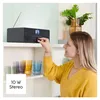 A woman is adjusting the volume on a black radio on a shelf, with colorful glasses below. Produktbild-Vorschau