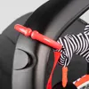 A close-up of a red clip attached to a black stroller handle, with a black and white striped toy hanging from it. Podgląd zdjęcia produktu