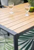 A close-up of a wooden table with a dark frame, set on a rug with a palm leaf pattern. Two glasses of white wine are on the table. Produktbild-Vorschau