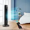 A person is holding a remote control in front of a tower fan in a bedroom. Voir les images du produit