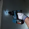A person wearing a protective glove is using a Bosch rotary hammer drill to make a hole in a concrete wall. The drill has a battery pack attached and a light illuminates the drilling area. Produktbild-Vorschau