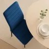 A blue velvet dining chair is placed next to a round wooden table with a place setting and a small vase of greenery. Produktbild-Vorschau