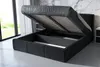 A modern black upholstered bed frame with the mattress and base lifted to reveal the storage compartment underneath. The bed is set in a well-lit room with a marble-patterned wall and a large window. Produktbild-Vorschau