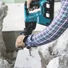 A person wearing a plaid shirt and work gloves is using a teal and black Makita rotary hammer to break apart a concrete block. Produktbild-Vorschau