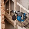 A person is using a blue and black rotary hammer drill to break apart a brick wall next to a wooden staircase. Podgląd zdjęcia produktu