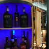 A wooden barrel-shaped cabinet displaying several bottles of alcohol, illuminated by blue LED lights. Produktbild-Vorschau