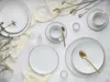 A flat lay of white porcelain dinnerware with gold trim, accented by dried flowers and small candles. Produktbild-Vorschau