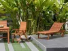 A wooden garden lounger and chair are set up on a patio next to lush green plants. Anteprima dell’immagine prodotto