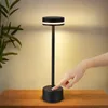 A hand is touching the base of a black LED table lamp on a wooden surface, with a plant in the background. Náhľad obrázku produktu