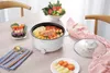 A white electric pot filled with food is placed on a wooden table, surrounded by various food items and cutlery, suggesting a meal preparation or serving scene. Produktbild-Vorschau