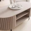 A close-up of a modern, oval-shaped coffee table with a built-in shelf and fluted side panels, placed on a light-colored carpet. Produktbild-Vorschau