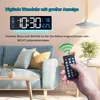 A digital wall clock is displayed on a wall above a sofa, with a hand holding a remote control in the foreground. Produktbild-Vorschau