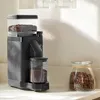 A black coffee grinder with a hopper full of coffee beans and a container of ground coffee sits on a countertop next to a glass jar of coffee beans. Produktbild-Vorschau
