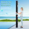 A woman is assembling a black outdoor shower by a swimming pool with the ocean in the background. Produktbild-Vorschau