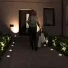 A woman walks up a lit pathway towards a house at night, carrying shopping bags. Produktbild-Vorschau