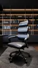 A black ergonomic office chair is placed on a dark rug in front of a bookshelf. Náhled obrázku produktu