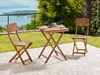 A wooden balcony set with two chairs and a table is placed on a paved patio overlooking a lake and greenery. Náhled obrázku produktu