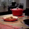 A red Dutch oven and a red frying pan filled with food are placed on a wooden table, with a checkered cloth and cutlery nearby. The background shows a blurred living room setting. Produktbild-Vorschau