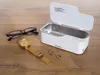 An ultrasonic cleaner is shown on a wooden surface next to a pair of glasses, a watch, and two rings. Voir les images du produit