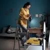 A woman is standing on a stepladder, holding a camera, in a room that appears to be a home studio. Náhled obrázku produktu