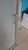 A close-up of a shower door handle and frame, with a frosted glass panel on the left and a white object and blue wall in the background. Produktbild-Vorschau