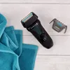 A black electric shaver with a blue trim and a grey trimmer attachment are laid out on a white wooden surface next to folded blue towels. Anteprima dell’immagine prodotto