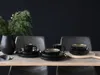 A table setting with black stoneware plates, bowls, and mugs, accented by a vase with greenery. Náhľad obrázku produktu