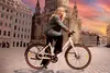 A woman rides an e-bike through a European city square with historic buildings and a sunset sky. Produktbild-Vorschau