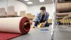 A man is kneeling on the floor, unrolling a red carpet and using yellow tape to mark a line. Náhled obrázku produktu