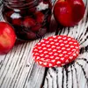 A close-up of a jar of preserves with a red lid patterned with white hearts, next to two red apples on a distressed wooden surface. Produktbild-Vorschau