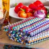 A stack of colorful kitchen towels in the foreground, with a bowl of vegetables and a glass of liquid in the background. Produktbild-Vorschau