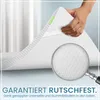A hand is holding up a corner of a white mattress topper, showing its textured underside and elastic corner straps. A magnifying glass is placed over the topper to highlight its texture. Text is present at the bottom of the image. Produktbild-Vorschau