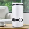 A white humidifier with a black accent and control knob sits on a wooden table next to a white coffee cup and saucer. In the background, a blurred view of a living room with a white sofa and a window with greenery is visible. Voir les images du produit