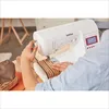 A person is using a white Brother sewing machine to sew a striped fabric with text on it. Produktbild-Vorschau