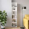 A white bookshelf filled with books and decorative items, placed next to a large potted plant and a yellow sofa with a floor lamp. Produktbild-Vorschau
