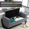 A grey upholstered storage bench with its lid open, revealing pillows inside. The bench is placed in a bedroom or living room setting. Podgląd zdjęcia produktu