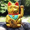 A golden Maneki Neko cat figurine sits on rocks outdoors with a blurred green background. Produktbild-Vorschau