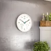 A white wall clock with thermometer and hygrometer is mounted on a textured gray wall. To the right, a white mantelpiece holds decorative items including bottles and a wooden box. A green plant is visible in the foreground. Voir les images du produit