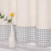 A white vase with a yellow flower and a green stem sits on a white surface in front of a curtain with a beige top and a gray checkered bottom. Produktbild-Vorschau