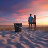 A couple stands on a sandy beach at sunset, looking out at the ocean. A dark gray bucket sits in the sand to their left. Anteprima dell’immagine prodotto