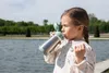 A young girl drinks from a metal straw cup by a body of water. Podgląd zdjęcia produktu