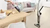 A person is using a handheld vacuum cleaner to clean crumbs off a wooden table. A standing vacuum cleaner is visible in the background. Produktbild-Vorschau