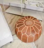 A brown leather pouf with white stitching sits on a rug next to a white sofa, with a shelf unit in the background. Produktbild-Vorschau