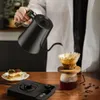 A person is pouring hot water from a black gooseneck kettle into a pour-over coffee maker, with a slice of cake and coffee beans on the table. Produktbild-Vorschau