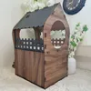 A wooden cat house with a dark roof and a small fence detail, placed on a white rug with decorative white flowers in the background. Voir les images du produit