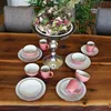 A table setting with pink and white ceramic dishes, a floral centerpiece, and chairs in the background. Produktbild-Vorschau