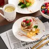 A table setting with breakfast items, including pancakes with berries, pasta with broccoli, a bowl of fruit salad, and a mug of tea, arranged on marble-patterned plates. Náhled obrázku produktu